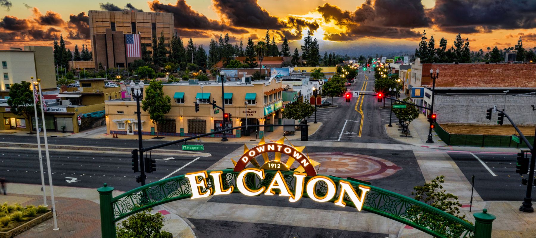 An aerial view of downtown El Cajon at sunset captures a prominent archway sign over a street, with buildings and streetlights illuminated by the warm sky