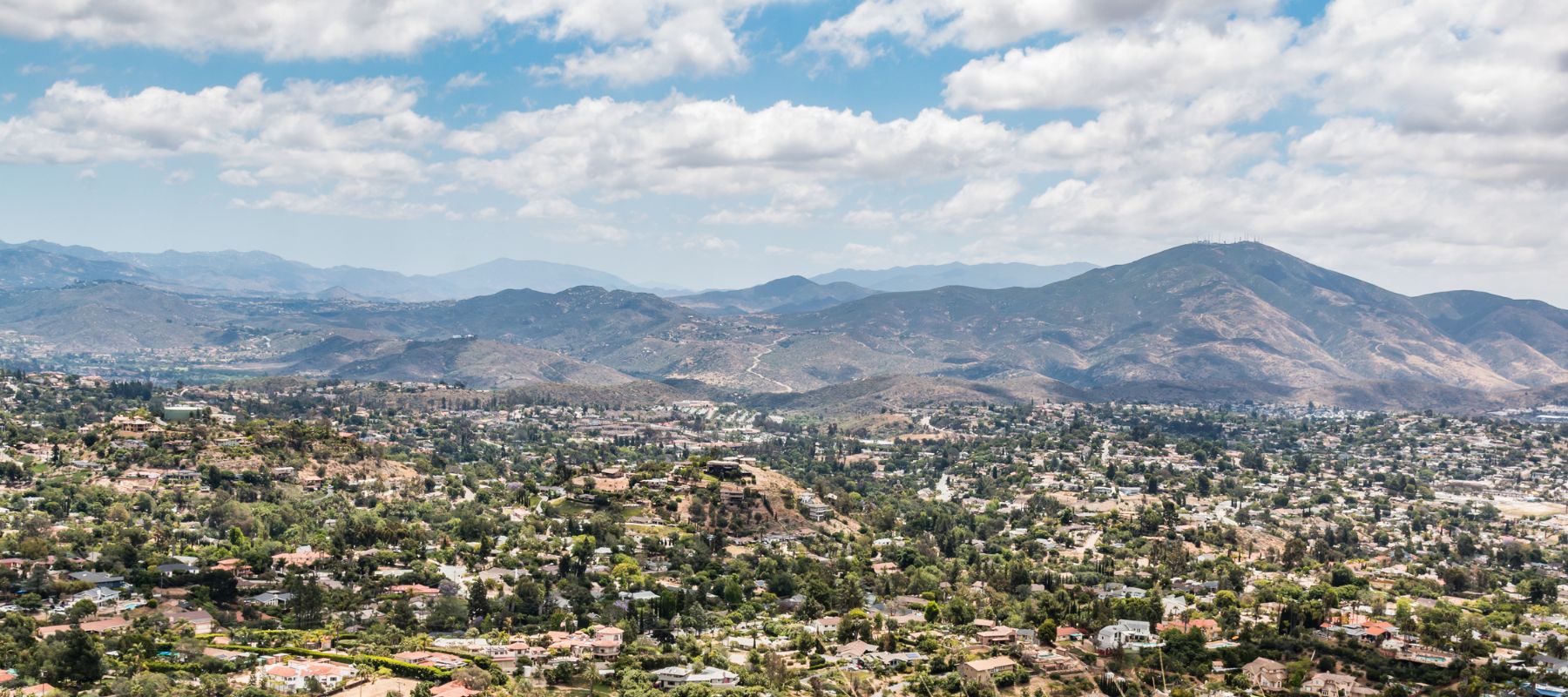 An expansive aerial view showcases a densely populated hillside residential area with green trees, extending towards distant mountains under a cloudy sky