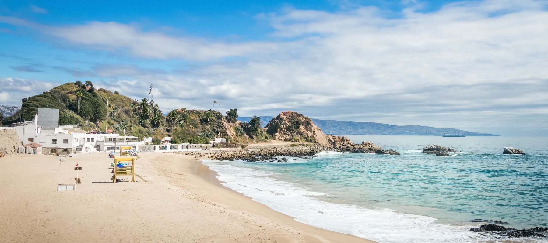 A sandy beach with gentle waves, clear blue water, and rocky cliffs in the background under a partly cloudy sky. There are a few buildings and beach structures along the shore.