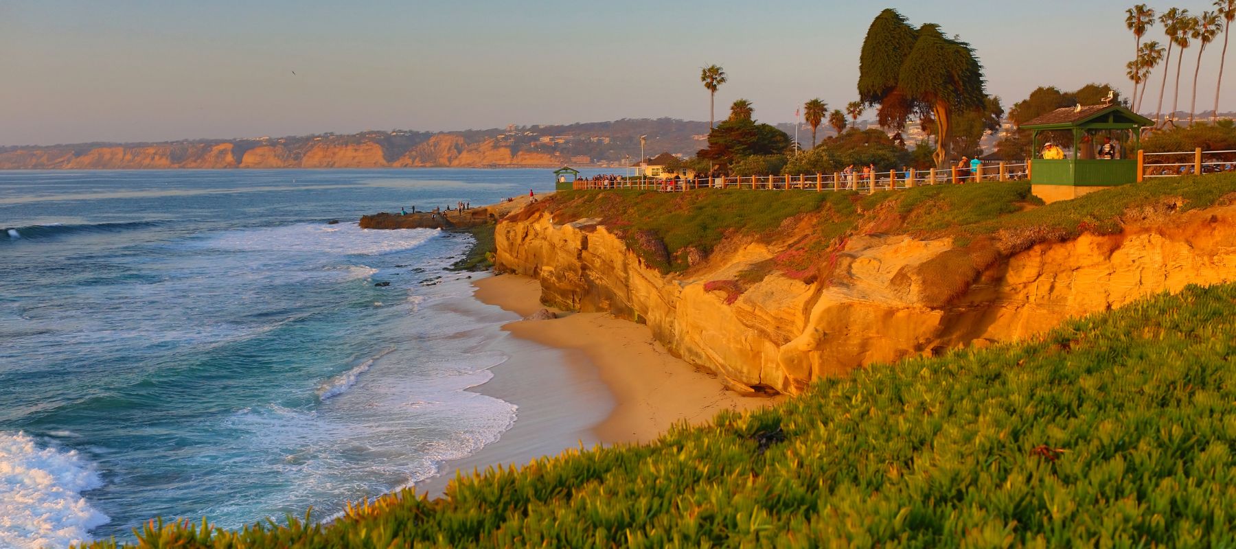 Golden cliffs overlooking the ocean at sunset, with waves crashing on a sandy beach below, palm trees, green shrubbery, and people walking along a fence-lined path atop the cliffs.