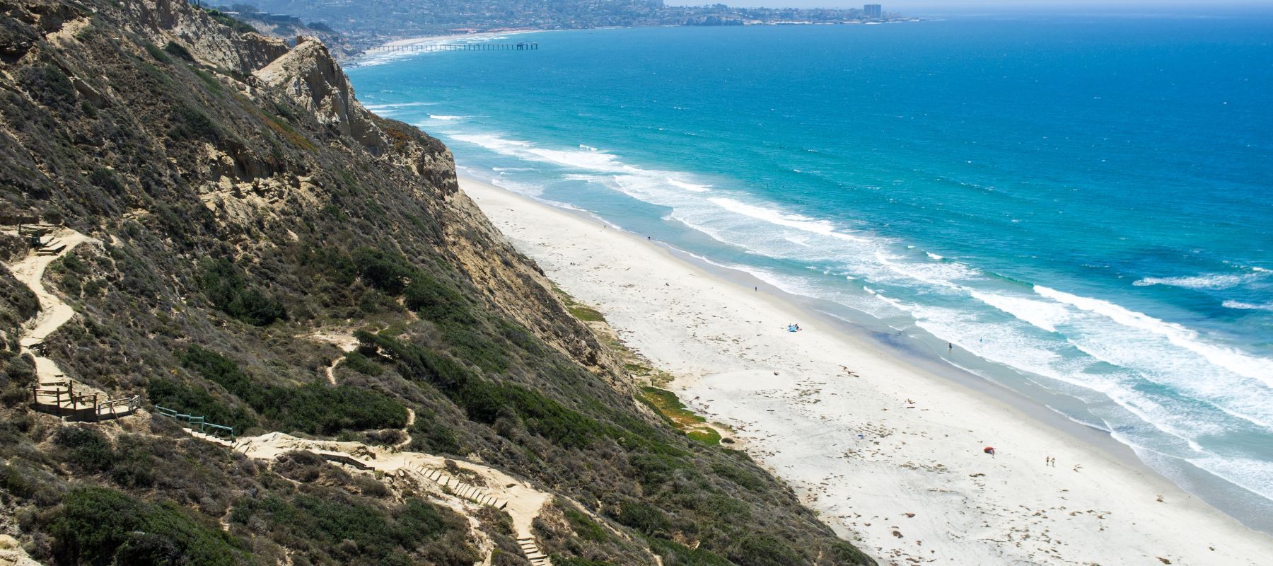 A view of a sandy beach bordered by blue ocean waves and rugged cliffs, with trails winding up the cliffside and a distant city visible along the coastline under a clear sky.