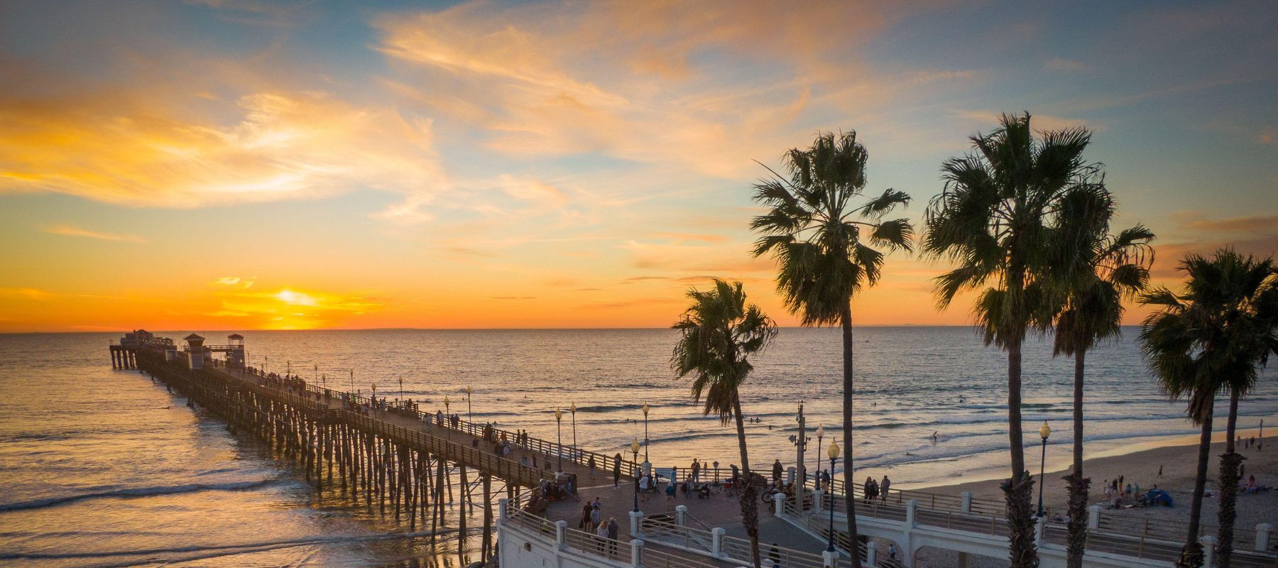 A long pier stretches into the ocean at sunset, with people walking along it. Palm trees and a crowd are in the foreground, and the sky is filled with colorful clouds over the water.