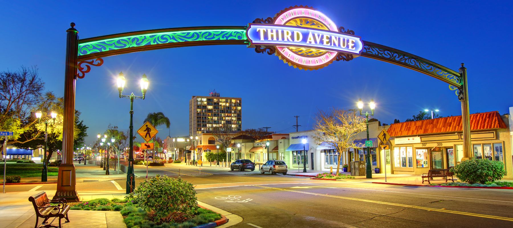 A brightly lit arch reading Third Avenue spans across a quiet street lined with shops, trees, lampposts, and benches at dusk, with a few cars and a tall building visible in the background.