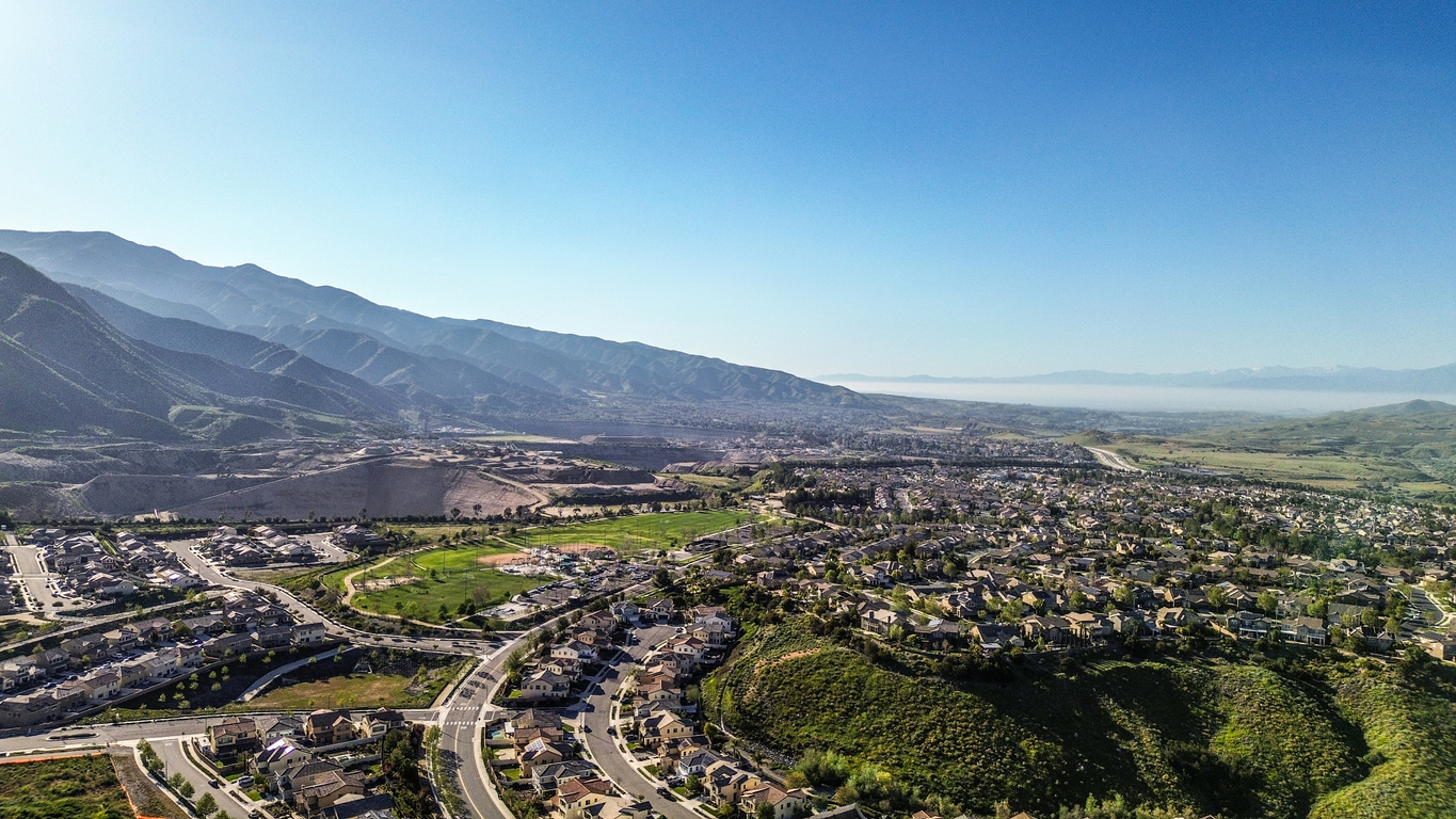 Aerial view of a suburban neighborhood with winding streets, green spaces, and houses, set against a backdrop of mountains under a clear blue sky.
