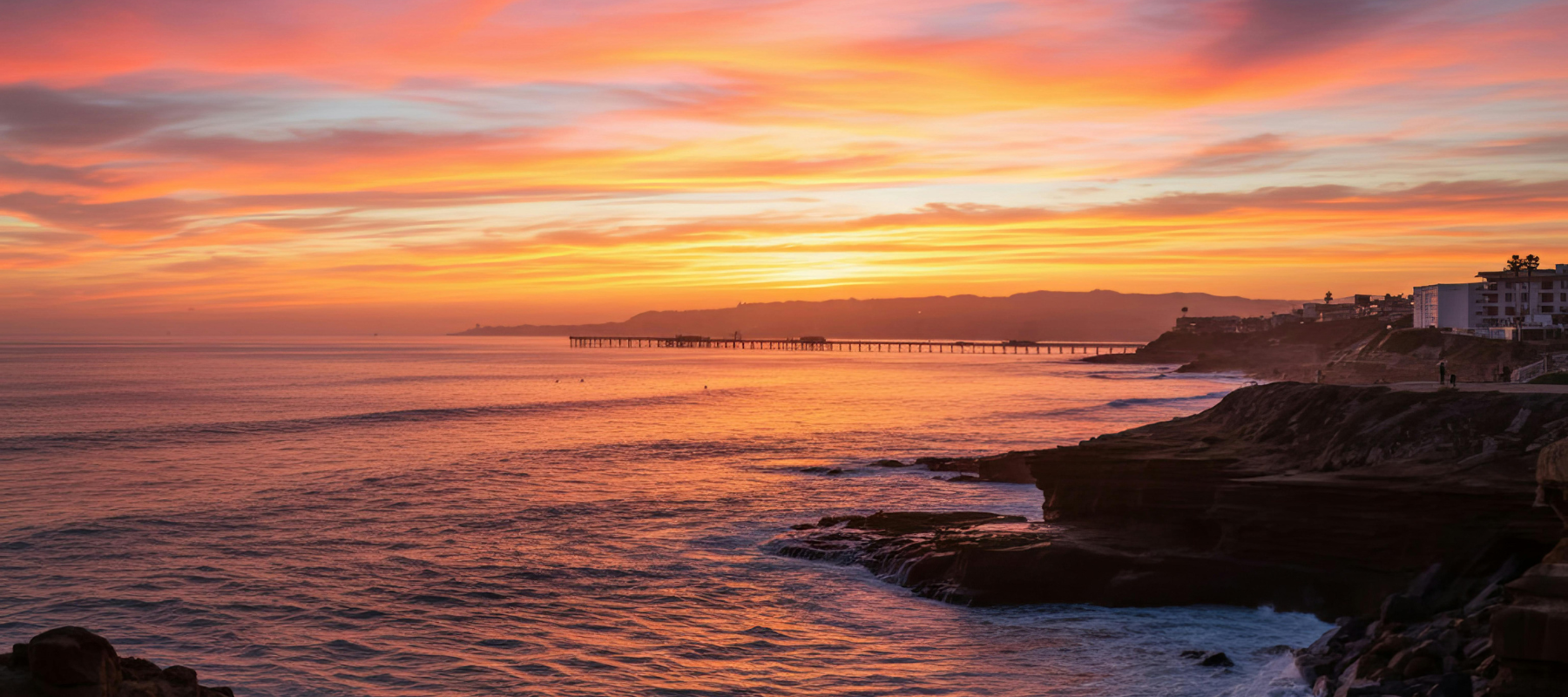 A vibrant sunset over the ocean, with colorful clouds in the sky, a rocky shoreline in the foreground, and a long pier stretching out into the water near distant buildings on the right.