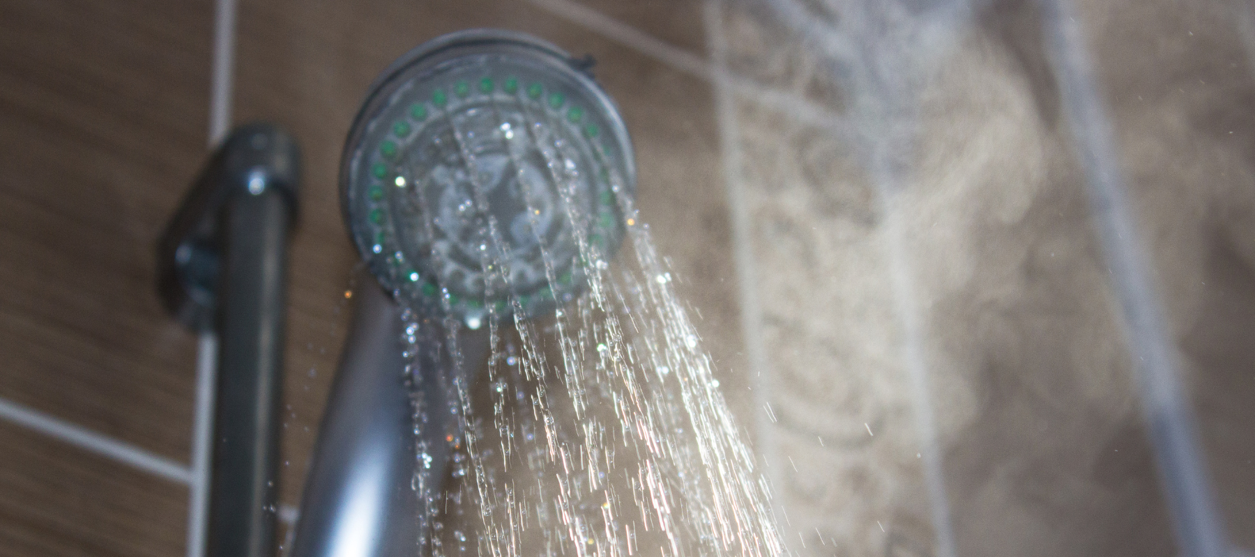 A close-up of a showerhead spraying water with steam rising, set against brown tiled walls in a bathroom.